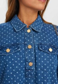 Chemise en denim avec un motif à pois, un col, des boutons dorés et deux poches avant avec rabats. Couleur bleue avec des textures variées.