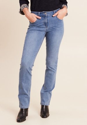 Frau trägt hellblaue, gerade geschnittene Jeans mit Händen in den Taschen, kombiniert mit schwarzen Stiefeletten mit Absatz und einem dunklen Oberteil.