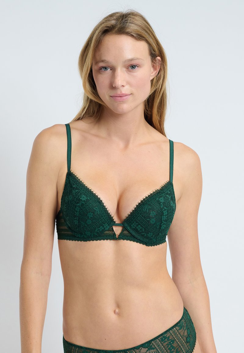 Reggiseno in pizzo verde smeraldo con design triangolare e imbottito; presenta spalline regolabili, una chiusura decorativa a gancio e intricati motivi floreali.