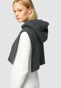 Una donna bionda che indossa una maglietta bianca a maniche lunghe e una sciarpa a cappuccio grigio scuro in maglia, appoggiata sulle spalle, mentre guarda di lato.