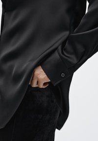 Black satin shirt with a smooth texture, featuring a buttoned cuff and clean tailoring. The shirt is tucked into black textured pants.