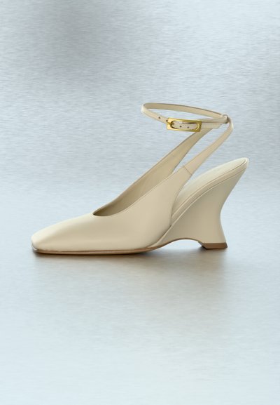 Κρεμ δερμάτινο παπούτσι slingback με χρυσή αγκράφα στον αστράγαλο και μοναδικά καμπυλωτό τακούνι πλατφόρμας πάνω σε λείο γκρι ανοιχτό φόντο.