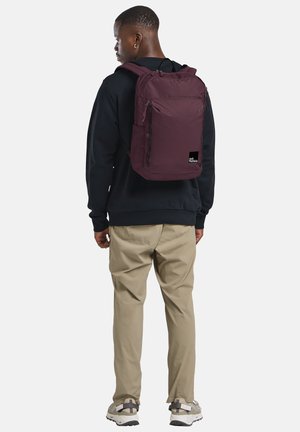 Bordeauxkleurige rugzak met twee ritscompartimenten, stoffen schouderbanden en een klein logo-patch aan de onderzijde van de voorkant. Geribbelde stof.