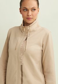 Donna che indossa una giacca beige con zip e colletto alto, con stringhe, mentre guarda di lato su uno sfondo chiaro e semplice.