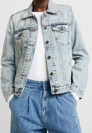 Personne portant une veste en jean bleu clair par-dessus une chemise blanche et un jean bleu ample, les mains dans les poches, sur un fond uni.