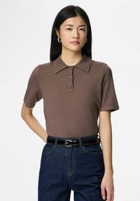 Brūna īsroku polo krekls ar teksturētu virsmu, trīs pogu aizdari un plakanu apkakli, kopā ar tumšiem džinsa džinsiem un melnu jostu.