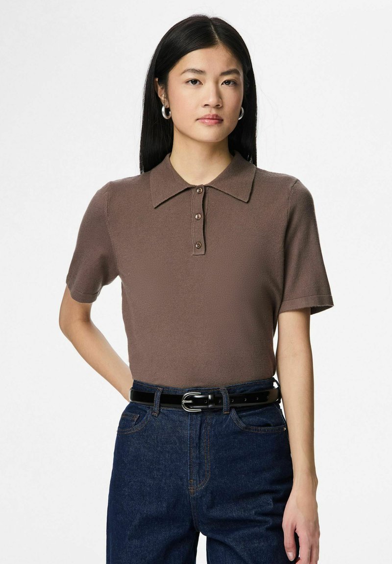 Brūna īsroku polo krekls ar teksturētu virsmu, trīs pogu aizdari un plakanu apkakli, kopā ar tumšiem džinsa džinsiem un melnu jostu.