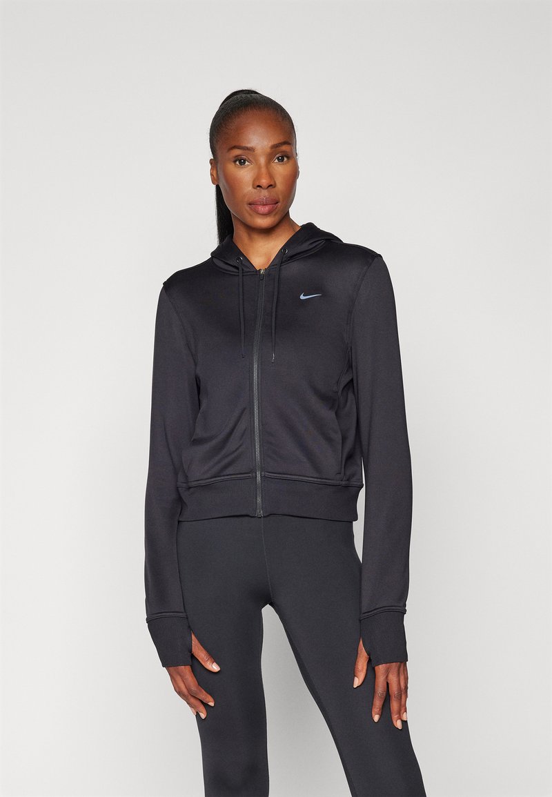 Juodas Nike zip-up džemperis su prigludusia siluetu, ilgomis rankovėmis ir ribotais manžetais. Yra mažas mėlynas logotipas kairėje krūtinės pusėje.