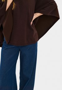 Rjav oversized top z širokimi rokavi, v kombinaciji z modrimi širokimi hlačami. Top ima gladko teksturo in sproščen kroj.