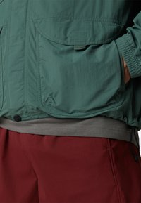 Grüne leichtejacke mit elastischen Bündchen, zwei Fronttaschen, über einem grauen Shirt und roten Hosen geschichtet, mit einer strukturierten Oberfläche.