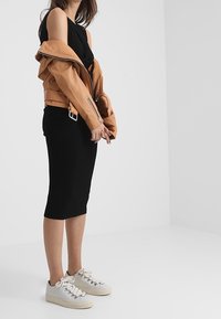 Femme portant une robe noire sans manches et des baskets blanches, en train d'enfiler partiellement une veste en cuir beige sur un fond blanc uni.