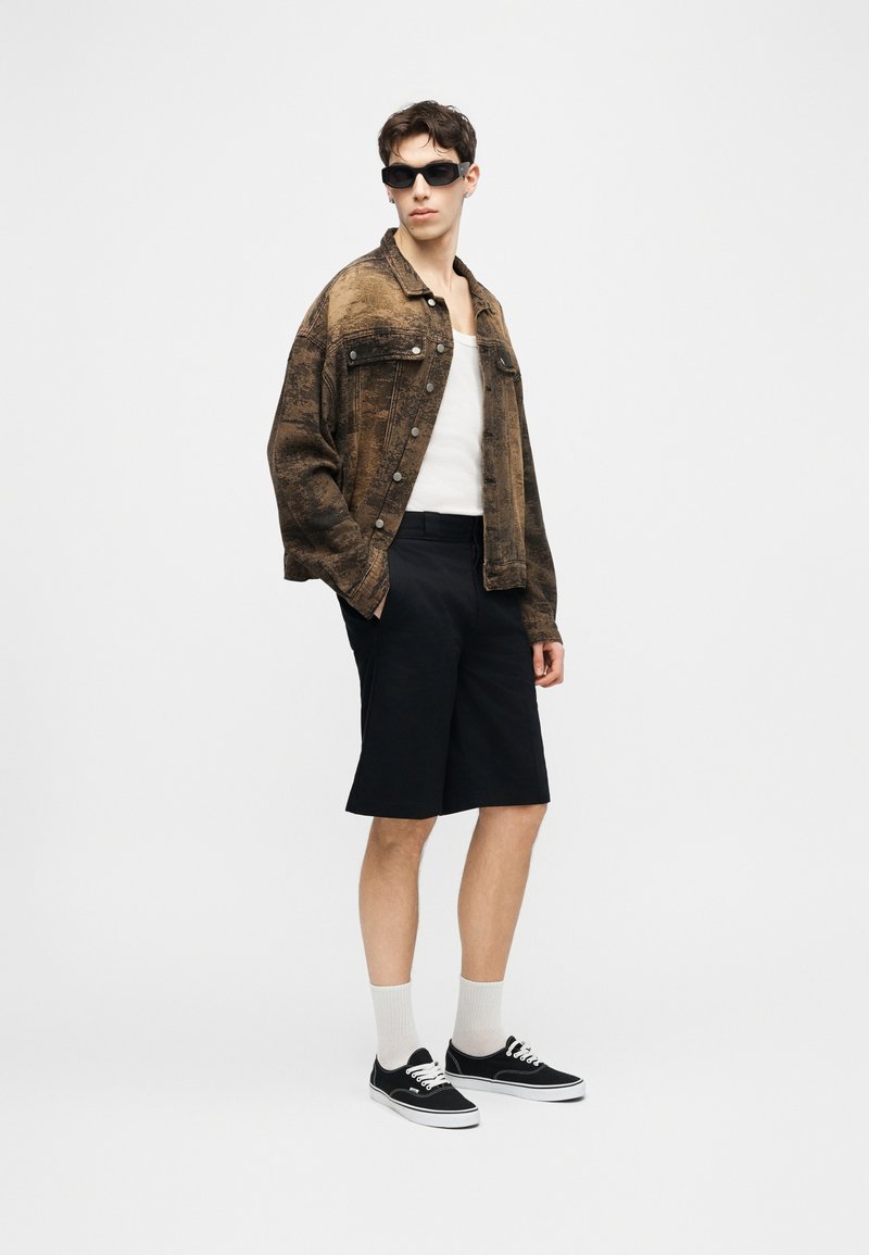 Giovane uomo con occhiali da sole neri, giacca marrone oversize, canottiera bianca, pantaloncini neri al ginocchio, calzini bianchi e scarpe da ginnastica nere, in piedi contro uno sfondo bianco.