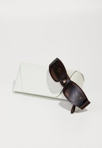 Ochelari de soare maro, supradimensionați, din plastic cu nuanță închisă, așezati pe un toc alb texturat cu text întărit care scrie "MICHAEL KORS".