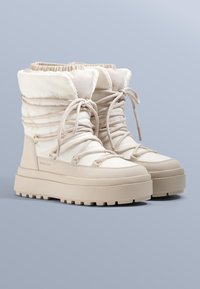 Bézs színű bélelt csizma párnázott felsőrésszel, kerek orral és vastag, tapadós gumitalppal, elöl fűzőrendszerrel rögzítve.