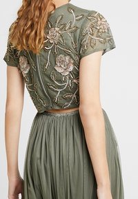 Groene crop top met korte mouwen, voorzien van opvallende bloem borduursels in metalen draden. Een vloeiende plissé rok maakt de outfit compleet.