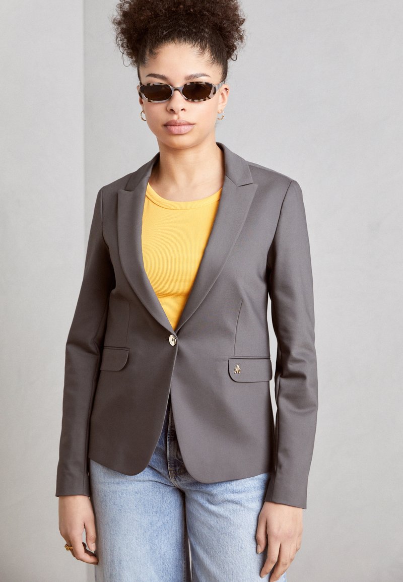 Šedý př tailored blazer s klopami, oboustranným zapínáním na jeden knoflík a dvěma předními kapsami, nošený přes žlutý žebrovaný top a světle modré džíny.