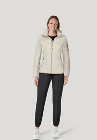 Lys beige zip-up jakke med højt krave, struktureret stof, sorte accenter, sidelommer; parret med mørke joggerbukser og hvide sneakers.