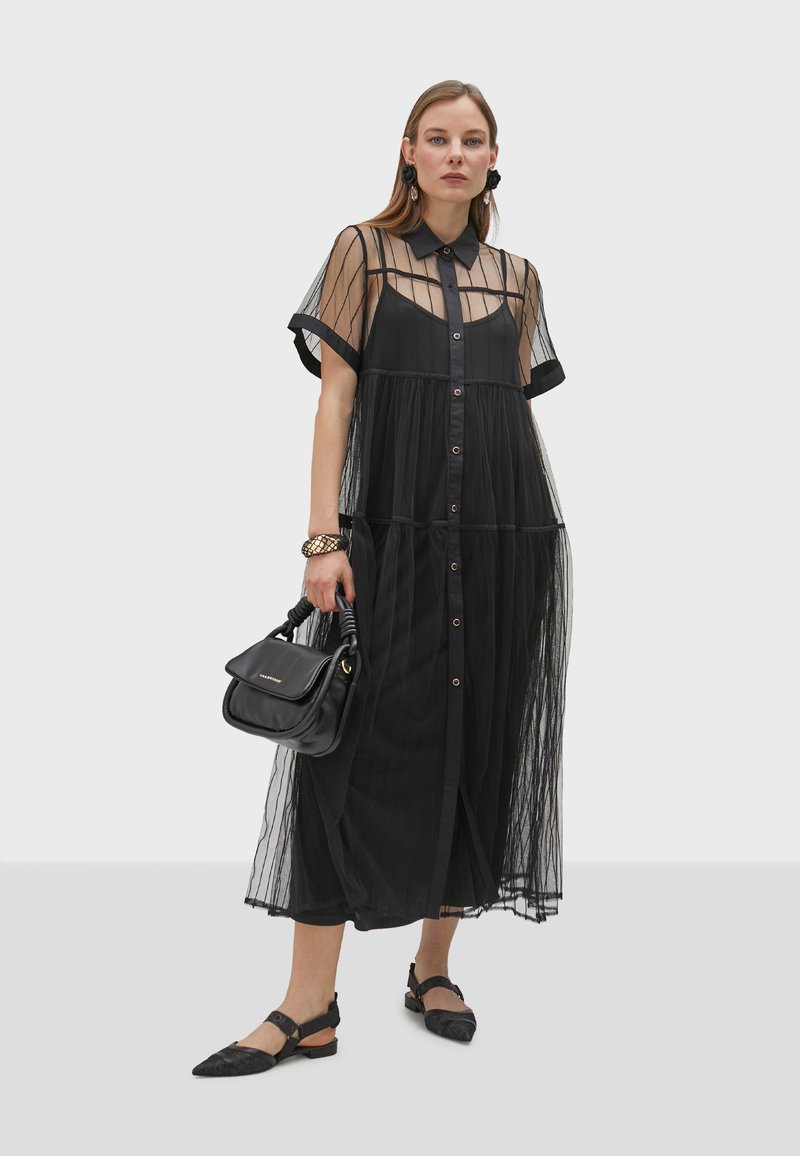 Robe noire avec superposition transparente, boutonnière à l'avant, manches courtes et tissu fluide. Associée à un sac à main noir et des chaussures plates.