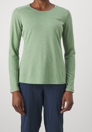 Personne portant un haut à manches longues vert clair avec le logo Vaude sur la poitrine et un pantalon bleu foncé, debout devant un fond uni.