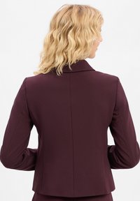 Kobieta z falującymi blond włosami, ubrana w dopasowaną ciemnofioletową marynarkę, pokazana od tyłu na białym tle.