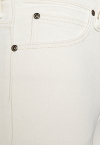 Primer plano de denim blanco mostrando un bolsillo con remaches metálicos negros, una textura sutil y costuras contrastantes a lo largo de las costuras.