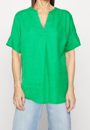 Frau trägt eine leuchtend grüne, kurzärmelige V-Ausschnitt-Bluse und hellblaue Jeans, steht vor einem schlichten weißen Hintergrund.