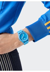 Sinine silikoonkell ümmarguse kellanäidikuga, minimalistliku disainiga, kontrastse tundide näitaja ja tekstuursete rihmacentidega. Kantakse randmel.