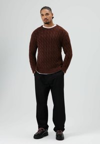 Gebreide bordeauxrode trui met kabelpatroon, gecombineerd met losse zwarte broek en zwarte schoenen. Voorzien van een ronde halslijn en een gestructureerde afwerking.