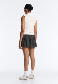 Femme debout, dos tourné, vêtue d'un top blanc côtelé sans manches, d'une mini-jupe plissée noire, de chaussettes blanches et de baskets beiges.