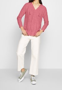 Femme portant un pull rose à manches longues avec col en V, un pantalon large blanc cassé et des baskets en toile vert clair, debout devant un fond uni.