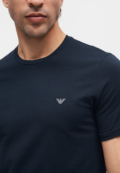 Άνδρας που φοράει μπλε σκούρο κοντομάνικο μπλουζάκι με μικρό γκρι στυλιζαρισμένο λογότυπο αετού στο αριστερό μέρος του στήθους.