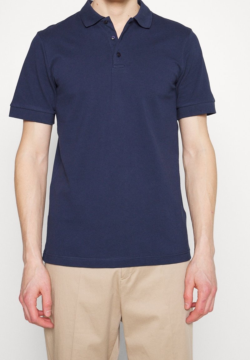 Homme portant un polo bleu marine uni à manches courtes et un pantalon beige, debout sur un fond blanc.