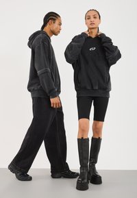 Svarte oversized hettegensere med ribbestrikkede mansjetter, sammen med mørke denimbukser og høye plattformstøvler til kneet. Glatte teksturer og minimale logoer.