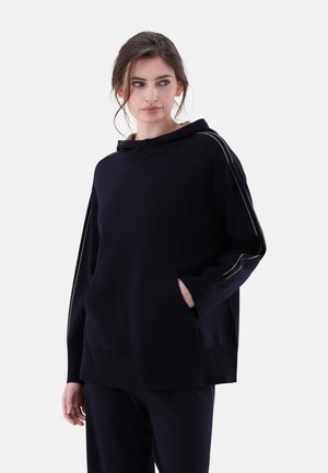 Felpa con cappuccio nera dal taglio morbido, con tessuto testurizzato, piping sui lati e tasca frontale. Abbinata a pantaloni scuri coordinati.