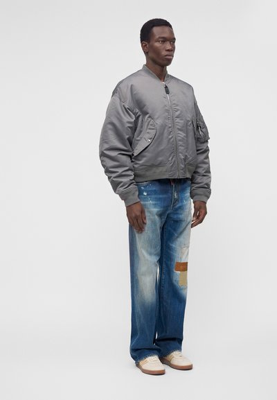 Homme debout portant une veste bomber grise, un jean bleu délavé avec des patchs et des baskets blanches sur un fond clair uni.