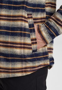Camicia di flanella con motivo a quadri navy, beige e ruggine. Presenta una tasca laterale e un tessuto texture. Maniche lunghe con dettagli ai polsini.