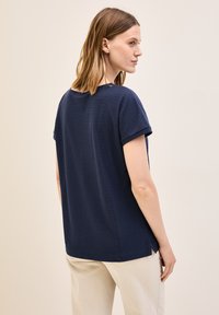 Camiseta texturizada de color azul marino con mangas cortas, dobladillo curvado y aberturas laterales, combinada con pantalones de color claro. Se muestra vista trasera.