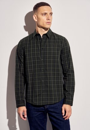 Chemise à manches longues de couleur vert foncé avec un motif à carreaux. Elle possède un col boutonné et une fermeture à boutons à l'avant, associée à un jean bleu.