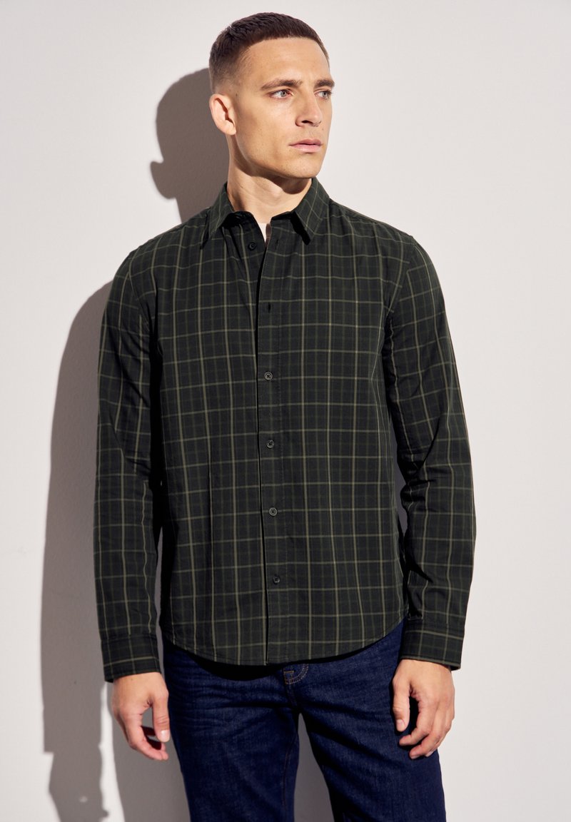 Hedvábná košile s dlouhými rukávy v tmavě zelené barvě s kostkovaným vzorem. Obsahuje límec s knoflíky a uzavření na knoflíky vepředu, kombinováno s modrými džínami.