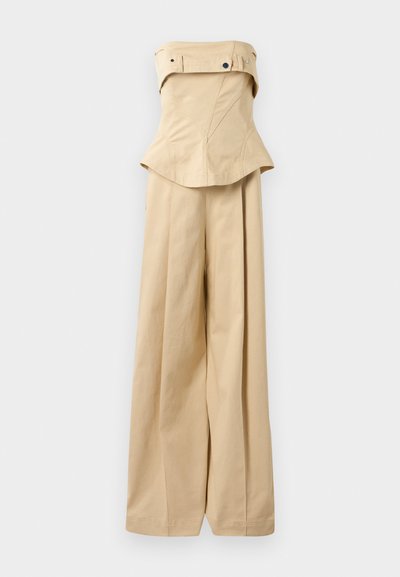 Macaquinho bege, sem alças, com um corpete estruturado, calças largas e um design peplum. Detalhes de botão e bolsos na parte superior.