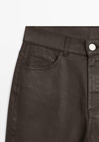 Detailní záběr tmavě hnědých kalhot se zobrazením přední kapsy, poutka na opasek, knoflíkové zapínání a detailního prošívání na bílém pozadí.