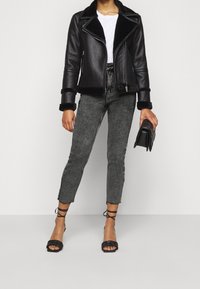 Chaqueta de cuero negra de motorista con cuello de borrego, combinada con jeans ajustados grises texturizados y sandalias de tacón negro con tiras. Cartera negra en la mano.