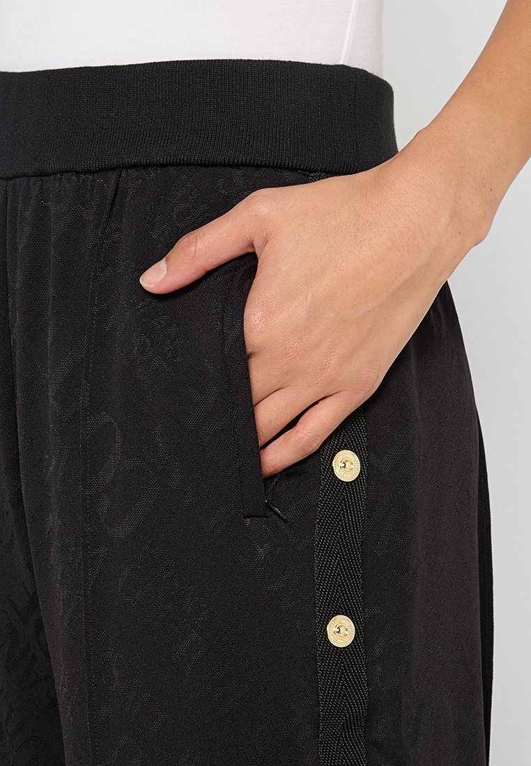 Schwarze Hose mit einem strukturierten Muster, ausgestattet mit einem breiten elastischen Bund und einer Seitentasche. Goldene Knöpfe akzentuieren die Seitennaht.