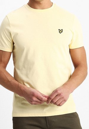 Homme portant un t-shirt jaune pâle à manches courtes avec un petit logo d'oiseau noir et jaune sur la poitrine gauche, mains jointes devant.