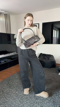 Mustan krokotiilikuviollisen käsilaukun pitkät kahvat, suorakaiteen muoto ja kiiltävä pinta, jonka henkilö pitää käsissään arkisissa harmaissa vaatteissa.