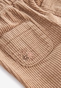 Tissu en velours côtelé marron clair avec des rainures verticales et une petite poche ornée de fleurs brodées discrètes en tons roses et bruns.