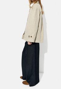 Beige oversized jakke med en afslappet pasform, som har store forreste knapper og sidelommer, parret med mørke vide denimbukser og brune slip-on sko.