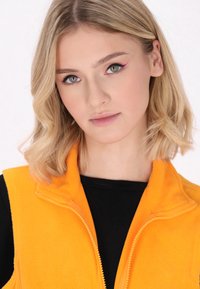 Gilet in fleece arancione con chiusura a zip, caratterizzato da un colletto alto. Il modello indossa una maglietta nera sotto. Texture liscia e colore vivace.