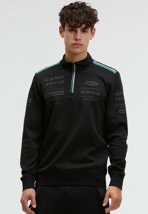 Junger Mann trägt einen schwarzen Reißverschluss-Pullover mit rennsportbezogenen Schriftzügen und hellblauen Streifen auf den Schultern vor einem einfarbigen Hintergrund.