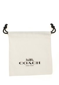 Valkoinen kanvasnauhatasku, jossa on musta painatus "COACH"-logosta ja vaunusuunnittelusta. Mustat naruinnit ylhäällä.